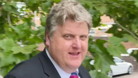 BBC Close of up Gregory Hill. He is wearing a white shirt, dark jacket and striped tie. He has wavy gray hair and a double chin