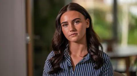 A young women with long wavy dark hair stares expressionless at the camera, wearing a blue and white striped shirt.