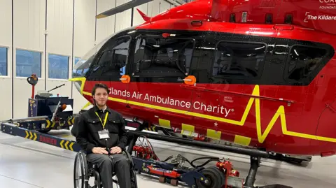 A young man with brown hair is sitting in a wheelchair. Next to him is a large red air ambulance with black tinted windows. On its bodywork there is a yellow line in the shape of a heartbeat from a monitor. In white writing it reads "Midlands Air Ambulance charity"