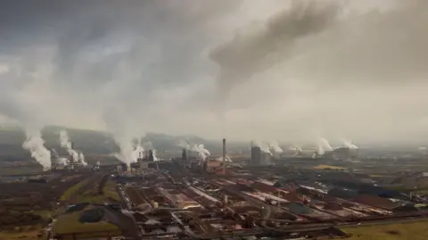 Matthew Harwood/Getty Images A panoramic view of the massive Port Talbot Steelworks shows numerous plumes of smoke from various structures against a gray sky.