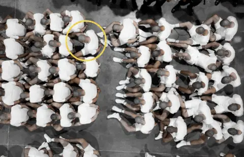 Una imagen de prisioneros sentados en un piso con calcetines blancos y pantalones cortos y camisetas blancas. Sus cabezas afeitadas se inclinan para que no veamos sus caras. Un círculo amarillo señala al señor García