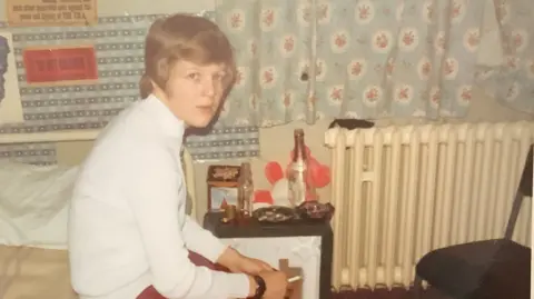 Pam Johnson A young woman with short hair sits on a bed, smoking a cigarette. The picture is taken in the 70s, so it looks old, and in the background you can see an empty bottle on a bedside table. The pattern on the curtains and the posters on th wall give the impression it is a teenager's bedroom.