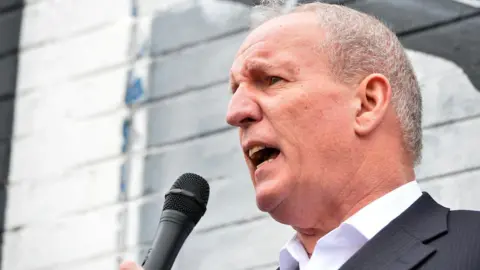 Bobby Storey is speaking into a mic. He's outside. A painted wall is behind him. He's wearing a black suit with a white shirt. 