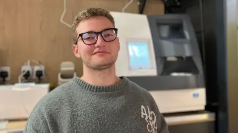 Steve Jones/BBC Vinnie Chambers, who has black-rimmed glasses and short blonde hair. He is looking straight at the camera and wearing a grey jumper.