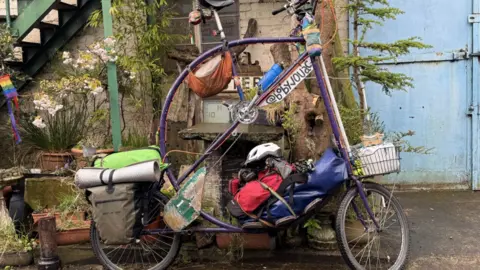 The bicycle has two small wheels and a large circle frame in the middle. It has been painted purple. It has a basket in the front with a water dispenser. There are a number of large backs attached onto the bike carrying a tent and other essential items. 