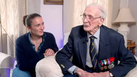 Vicki McClure on the left wearing a dark-coloured jumper and sitting on the floor next to her grandfather Ralph. He is sat in an armchair wearing a navy blue suit, light-coloured shirt, navy blue tie with yellow diagonal stripes  and four war medals are pinned to his jacket.