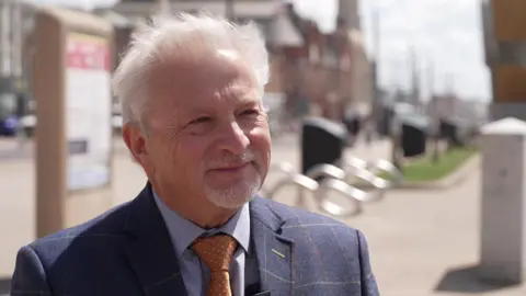 A picture of Alan Cavill taken on Blackpool Promenade, with the Tower in the background. He's wearing a blue suit and yellow tie and has white hair and a beard.