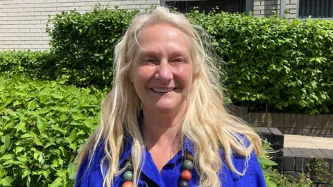 Head-and-shoulders image of Liz Crook standing outside in the sunshine in front of a green hedge. She has long blonde hair and is wearing a bright blue blouse and a long, multi-coloured chunky-beaded necklace.