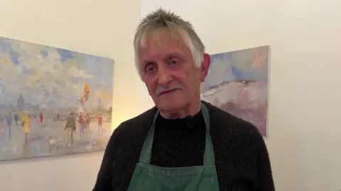 Stewart is wearing a black jumper with a dark green apron over it. Two paintings are on the walls behind him. One shows a scene with people in a square.