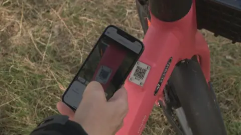 A man wearing dark clothing holding a phone which is scanning a QR code on a red bike frame.