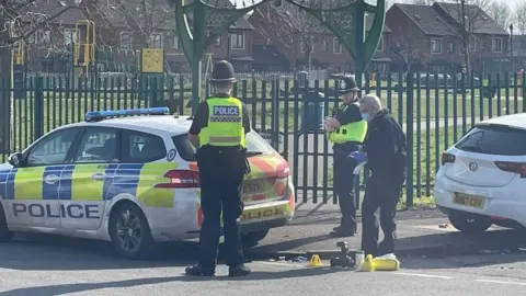 Three police officers stood near evidence markers and a police car