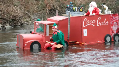 Sam Hepburn Two elves with oars sit at the front of a raft shaped like a red Coca-Cola lorry. A person dressed as Santa Claus sits at the top.