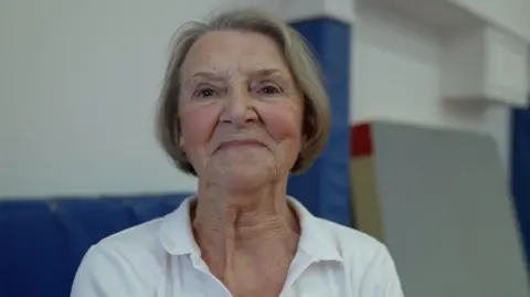 BBC Head and shoulder shot of octogenarian Anita Waters. She looks younger than her years, with light blonde bobbed hair and wearing a white open-necked shirt.
