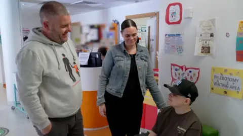 Ollie, sitting down in a hospital unit, wears a black baseball cap and a dark, round-necked top. A grey-haired man wearing a grey hoodie and a dark-haired woman wearing a blue denim jacket stand next to him.