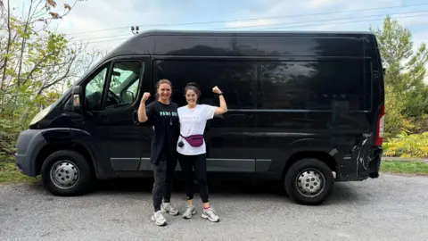 Kat Samuelson/EmpowerVan Kat Samuelson pictured with another volunteer from EmpowerVan. Both Kat and the other volunteer are holding one arm aloft in the picture. They are standing behind a black van. They are each wearing casual gym clothes.