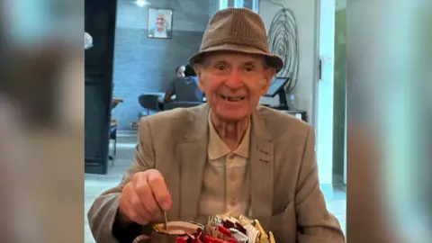 An elderly man smiles (right) for a photo in a cafe. He is wearing a pork-pie hat and a cream jacket and light shirt. He is stirring a hot drink in a mug on a table in front of him which contains a mental container of condiments