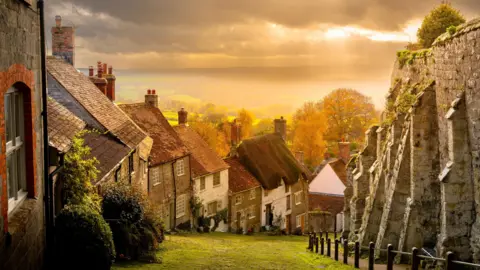 View from feet today Gold Hill in Shaftesbury. A row of houses on a steep hill. There is a large stone wall on one side. The sky is glowing orange with fields and hills behind.