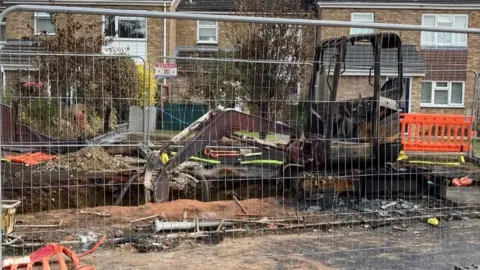 Rose Scott A metal fence surrounds a digger which has been burnt. There are houses in the background and several trees.