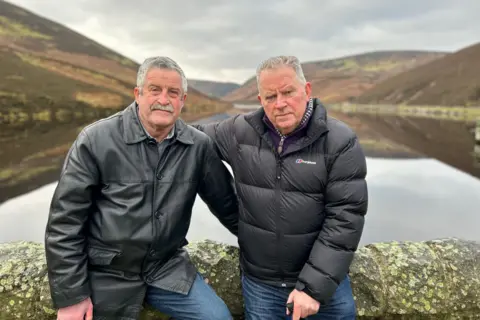 Mr Allan and Mr Anderson sitting on a wall with a reservoir behind them. They have their arms around each other.