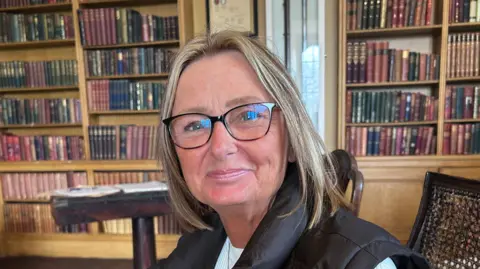 BBC/Kate Bradbrook Dawn Simpson-Lane, who has shoulder-length blonde hair and is wearing glasses, smiles at the camera. The walls behind her are lined with bookcases.