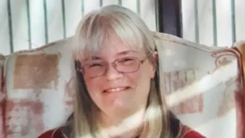 Handout Ms Fish pictured in a handout photo smiling and looking into the camera. She has got grey shoulder-length straight hair and a fringe, and is wearing glasses. She is sat in a large armchair with a pink and orange print, with a striped wall visible beyond it. 