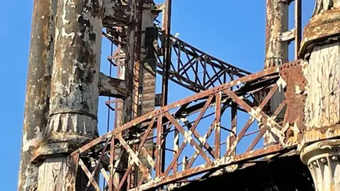 Andrew Turner/BBC The Great Yarmouth gasometer stands in a blue sky, with it's peeling paint and rusting of the columns and lattice work. This is a close up of the lattice work, and shows the gauge that once indicated the quantity of gas within, in cubic feet. With finials on the top it is is highly decorated, with it being awarded Grade II listed status.
