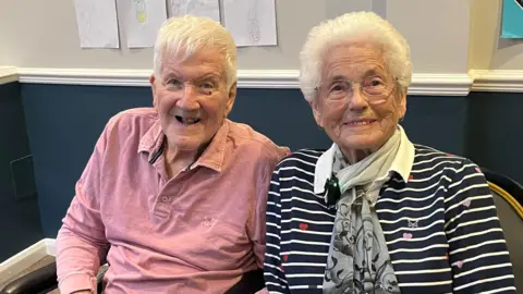 Bill and Olive Miles sat next to each other. Bill is wearing a pink shirt and Olive is wearing a striped navy and white shirt. Both Bill and Olive have white hair.