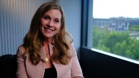 Julie Inman Grant in a pale pink jacket and black blouse smiles as she looks at the camera. She is sitting next to a window. 