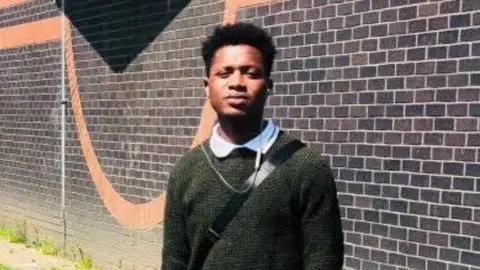 Johnson Ewere - a man with short dark hair and a green jumper, standing outside a redbrick building