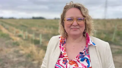 Stephen Huntley/BBC A woman in a field, by planted trees. She is wearing a cream jacket, floral shirt, with a necklace around her neck. She has short curly hair and is wearing glasses.