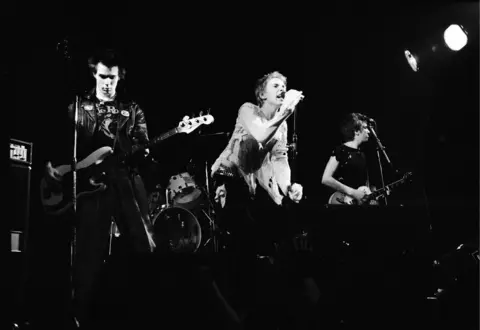 The Sex Pistols on stage in 1977. It is a black and white photo. Johnny Rotten is seen snarling into a microphone in cut up-clothing.  Sid Vicious wears a leather jacket while Steve Jones is in a crop black top. 