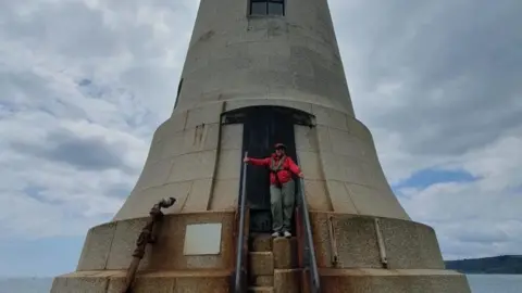 Jennifer Kloester to the right wearing a red coat, red cap and dark green trousers. She is wearing sunglasses and is stood on one of the alternating steps going up to the Plymouth breakwater lighthouse door. The lighthouse is light grey and has a window at the centre.