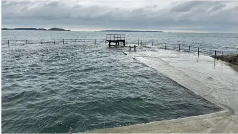 BBC The diving board is pictured at high tide against a grey sea and grey sky.