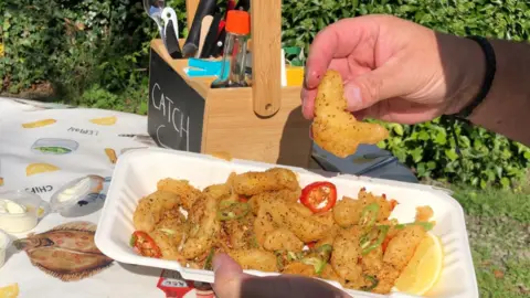 Stephen Peel A man's hand can be seen holding a chip with one hand and a container of chips with the other. The chips have herbs and cut up pieces of fresh chilli on them.