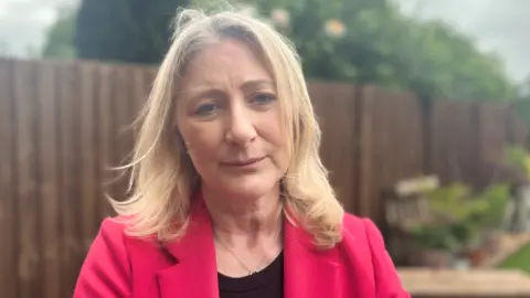 A woman with long blonde hair and a bright pink jacket standing in front of a fence in a garden