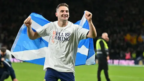 Getty Images Kenny McLean on the pitch holding a large Scottish flag behind his back. He is wearing a white shirt with the words “WE’LL BE COMING” and blue shorts with the number 23.