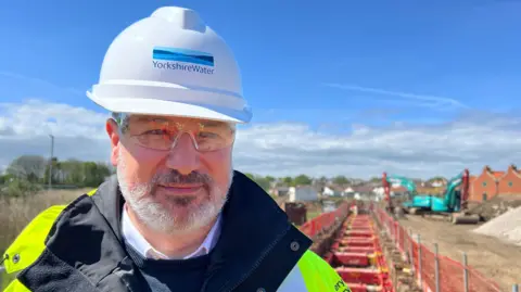 Miles Cameron from Yorkshire Water standing in front of a long trench. He wears a high vis jacket, a white hard hat with the Yorkshire Water logo on the front, and protective glasses. A work site is in the background. 