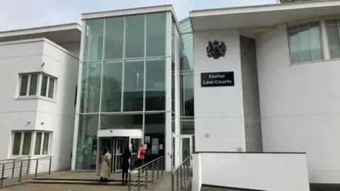 BBC Exterior of Exeter Law Courts featuring modern architecture with large glass panels and white walls. Three people are near the entrance, with one entering the building.