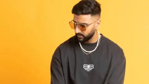 A man with short dark hair and a beard in a black top and sunglasses is standing in front of a yellow background.