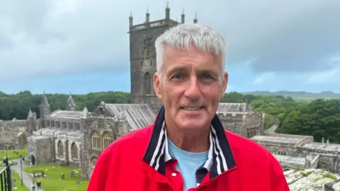 Iain smiling whilst standing in front of Cathedral. He wears a red jacket and a light blue t-shirt. He has short grey hair.