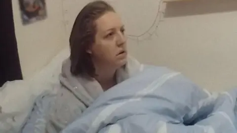 Netflix Police body-camera footage showing Lucy Letby, 35, who has blonde hair and is wearing a grey dressing gown, sitting upright in a bed with a light blue duvet around her. She is looking towards an officer with a shocked expression. 