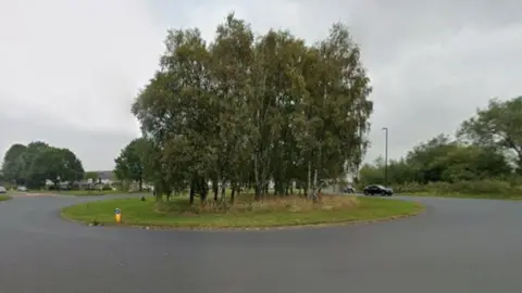 Google A roundabout with a cluster of tall, leafy trees planted in the centre on a grassy island. The road surrounding the roundabout is wide and paved, with a smooth, dark surface. Several streetlights are positioned around the roundabout, and a few vehicles can be seen in the background, including a black car approaching from the right side. The surrounding area appears to be open and green, with additional trees and shrubs visible beyond the roundabout. The sky is overcast.