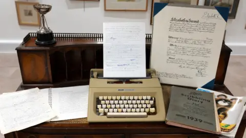 RWB Auctions A wooden desk displaying items from the private collection, including a silver golfing trophy, a vintage typewriter, a scribbling diary, and typewritten notes.