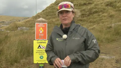 Ella Briens is wearing a dark green jacket and has sunglasses perched on the top of her hat. She is standing in moorland by a red sign which reads High fire risk, above another sign which says Camping is not permitted in this location.