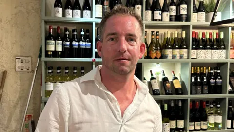 Pub landlord Tom Rowell, standing in front of a rack of wine bottles. He is wearing a white shirt and has short brown hair and stubble.