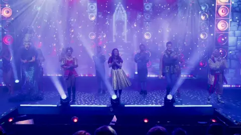 BBC A stage with blue lighting and about ten actors dressed as characters from Snow White, including Snow White in the middle and the dame on the far left 