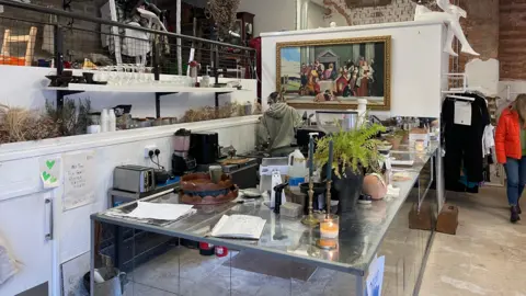 A general view of the café. There is a large mirrored counter space with lots of items on it, including a candlesticks and a plant, with a shop/gallery area behind it. Nathan is behind the counter wearing a hoody and facing away from the camera. There is a woman in a red coat to the right of the image perusing the rails.