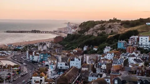 An aerial shot of a seaside town, with houses in the foreground and the sea - and a pier - in the distance