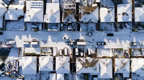 aerial view showing the tops of snow-clad roofs on houses and cars lining a street