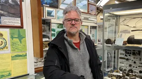 Shaun Whitmore/BBC Rob Spray, wearing a grey fleece and black jacket, is looking at the camera with a range of framed photos of World War Two planes behind him. He's standing in the Norfolk and Suffolk Aviation Museum near Bungay.